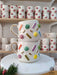 Handmade ceramic mugs with colorful candy-like sprinkles in various shapes, adorned with gold accents and a gold handle, stacked on a marble stand with festive greenery.