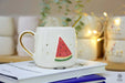 A handmade ceramic mug featuring a playful watermelon slice design with gold accents, placed on a book, perfect for a refreshing vibe.